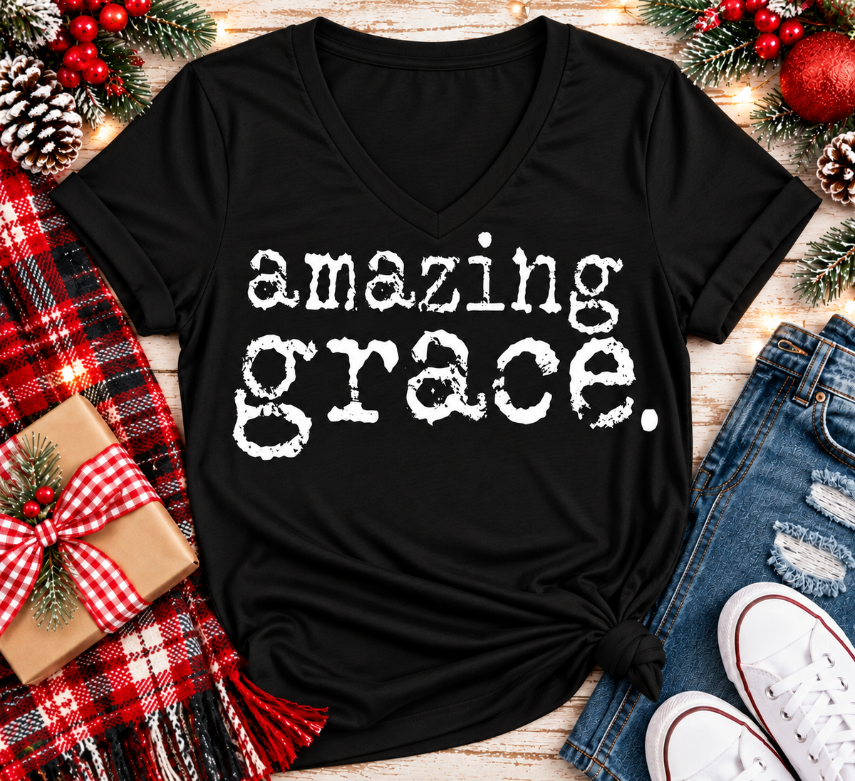 A black V-neck t-shirt with the words 'amazing grace.' printed in a distressed white typewriter font, styled in a flat lay with jeans, white sneakers, a red plaid scarf, a wrapped gift box, and Christmas pinecones and berries.
