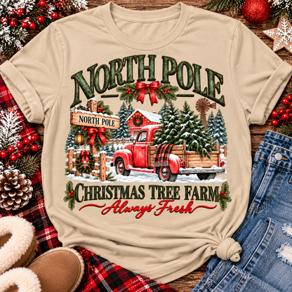 North Pole Christmas Tree Farm vintage holiday graphic shirt featuring a red pickup truck carrying Christmas trees, snowy evergreen trees, rustic farmhouse scenery, festive red bows, and distressed green lettering reading North Pole Christmas Tree Farm Always Fresh. Available in beige and black in short sleeve crewneck, V-neck, long sleeve crewneck, and hoodie styles.