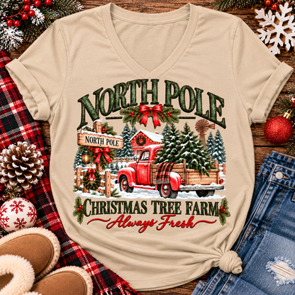 North Pole Christmas Tree Farm vintage holiday graphic shirt featuring a red pickup truck carrying Christmas trees, snowy evergreen trees, rustic farmhouse scenery, festive red bows, and distressed green lettering reading North Pole Christmas Tree Farm Always Fresh. Available in beige and black in short sleeve crewneck, V-neck, long sleeve crewneck, and hoodie styles.
